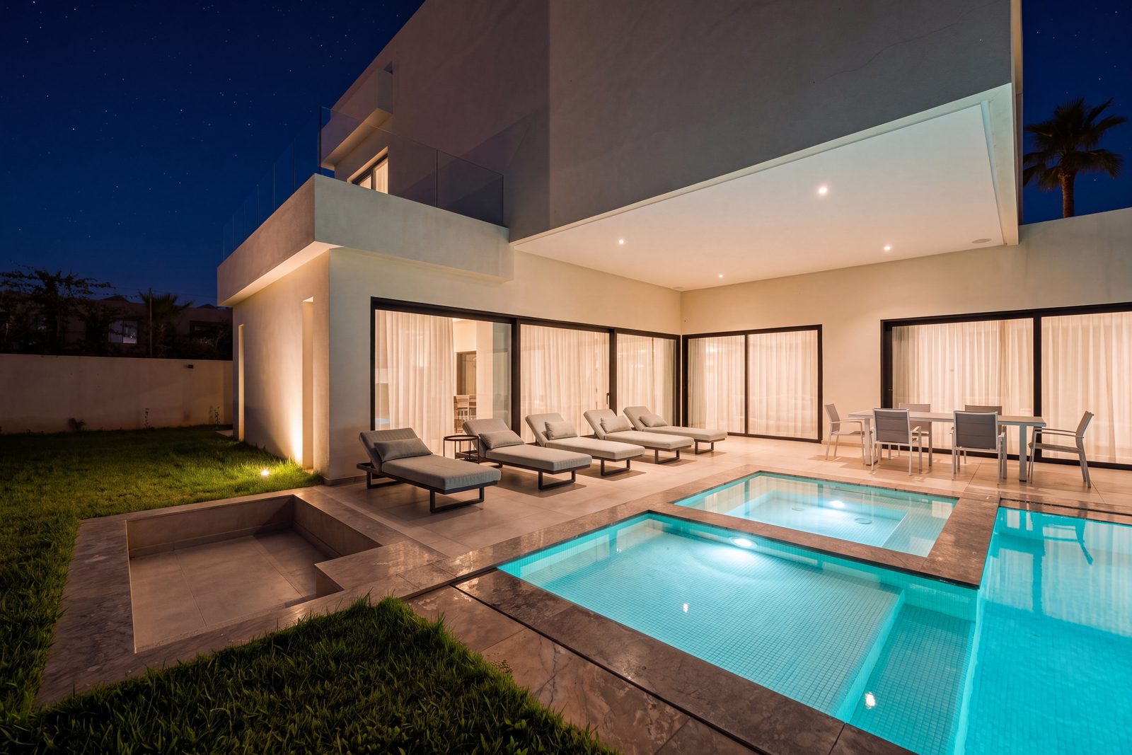 A modern villa at night: sun loungers and a dining table under a lit, curtained terrace, with a pool and a jacuzzi glowing in front.