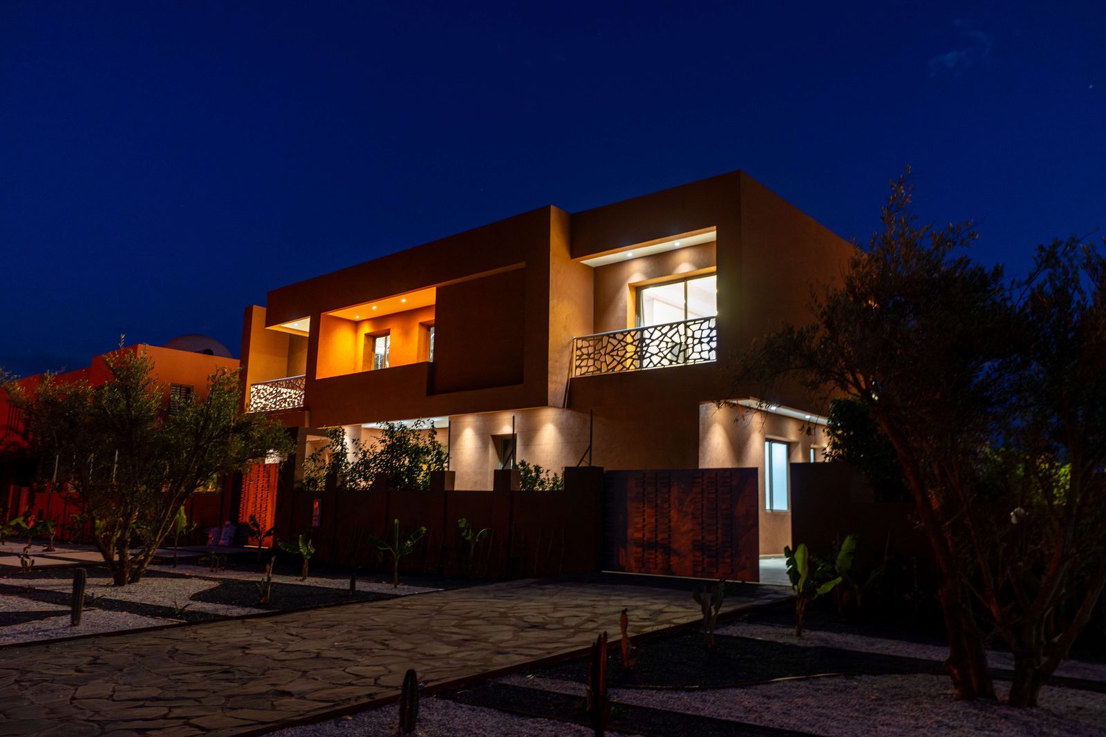 A villa at night: warm light behind the windows and balconies, a garden and palms in the foreground.